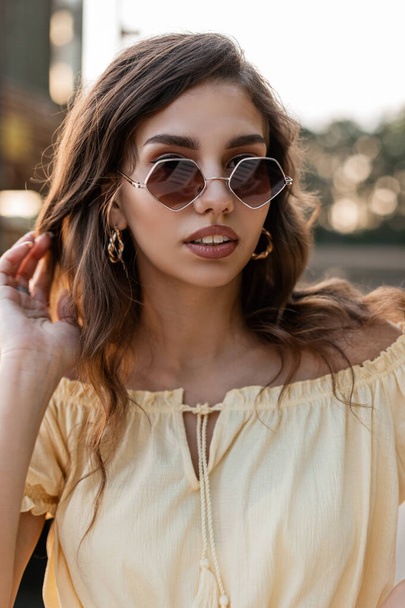 Ritratto fresco di attraente giovane donna riccia in camicia gialla alla moda con occhiali da sole vintage cammina all'aperto in una giornata di sole. - Foto, immagini