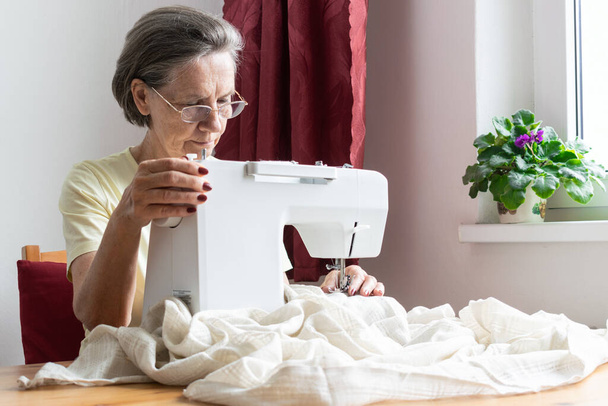 Ana konu odak dışı, yaşlı kadın kıdemli ev işi masasında oturuyor, dikiş makinesinin keyfini çıkarıyor rahatlıyor ve ev yapımı para kazanıyor. - Fotoğraf, Görsel
