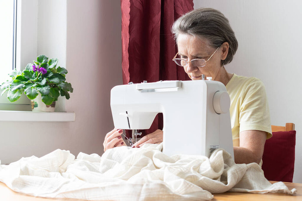 главная тема вне фокуса, старушка-старшая сидит рядом за столом за домашней работой, швейная машинка наслаждается счастливым отдыхом и зарабатывает деньги по дому - Фото, изображение