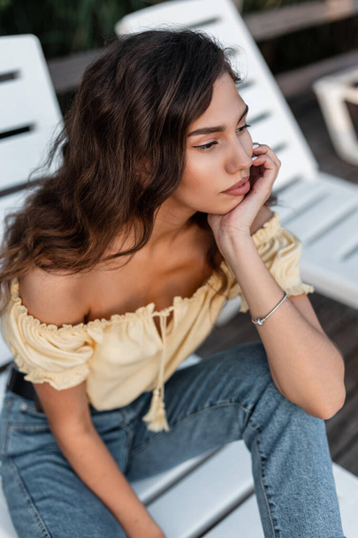 Pretty young woman with curly hair in blue jeans and yellow top blouse sits on the beach lounger - Fotoğraf, Görsel
