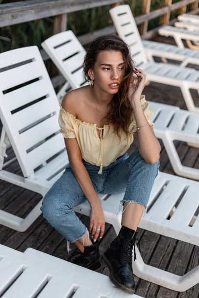Bella modella hipster donna in abiti di moda estate denim con camicetta gialla superiore, jeans blu e scarpe nere si siede e riposa su un lettino sulla spiaggia all'aperto - Foto, immagini
