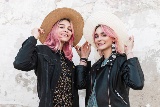 Mooie vrolijke jonge vrouwen met roze haar in modieuze kleding glimlachen en rechte stro stijlvolle hoeden in de buurt van witte muur in de stad. Charmante meisjes zussen poseren in de stad. Schoonheidsdames. - Foto, afbeelding