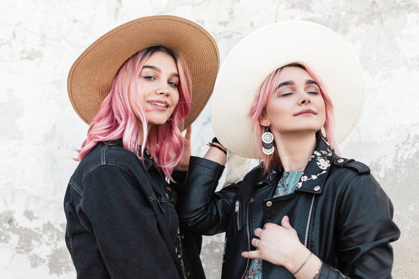 Δύο όμορφες αδελφές με χρωματιστά μαλλιά σε μοντέρνα ψάθινα καπέλα με κομψά casual σακάκια απολαμβάνουν διακοπές κοντά σε vintage λευκό κτίριο στο δρόμο. Μοντέρνα μοντέρνα όμορφα κορίτσια χαλαρώνουν σε εξωτερικούς χώρους. Καλοκαίρι. - Φωτογραφία, εικόνα