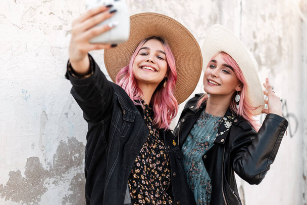 Две положительные молодые женщины с розовыми волосами в модной одежде в соломенных элегантных шляпах делают селфи на современной фотокамере возле белой стены на улице. Счастливые девушки улыбаются и сами фотографируют. - Фото, изображение
