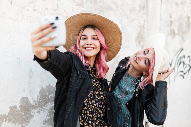 Two happy beautiful funny girls in fashionable denim clothes with a dress and hats take a selfie photo and enjoy a walk in the city - Foto, imagen