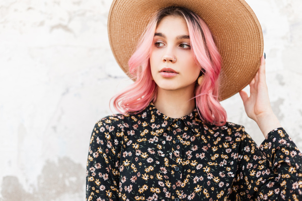 Красотка изображает симпатичную стильную молодую женщину в винтажной соломенной шляпе в элегантном красивом цветочном платье возле дома на улице. Милая девушка модель с розовыми волосами выпрямляет шляпу на открытом воздухе - Фото, изображение