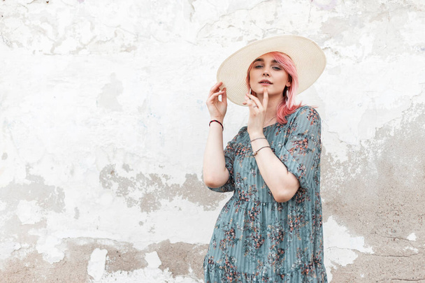 Pretty gorgeous young woman with chic pink hair in fashionable white  hat in summer stylish blue floral dress posing near wall on street. Sensual cute girl fashion model in trendy elegant wear in city - Fotoğraf, Görsel