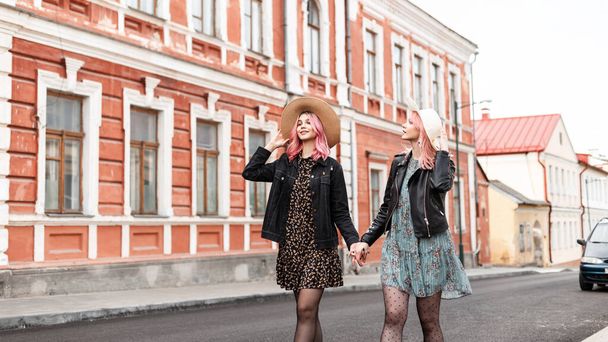 Αστική θετικά νεαρές γυναίκες φίλες στη μόδα κομψά ρούχα της νεολαίας σε κομψά μπουφάν σε vintage ψάθινα καπέλα απολαύσετε μια βόλτα στο δρόμο στην πόλη. Χαριτωμένα χαρούμενα κορίτσια με χαρούμενο χαμόγελο με ροζ μαλλιά ταξίδια. - Φωτογραφία, εικόνα