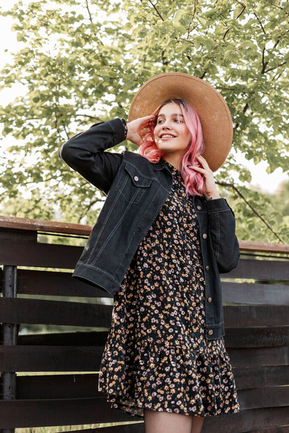 Dobrze uśmiechnięta młoda kobieta z kolorowymi różowymi włosami w słomkowym kapeluszu w stylowej kurtce jeansowej w sukience modowej relaksuje się w pobliżu drewnianego ogrodzenia na wsi. Szczęśliwa dziewczyna ze słodkim uśmiechem cieszy się odpoczynkiem na łonie natury. - Zdjęcie, obraz