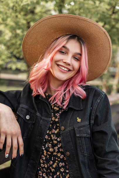 Cute smiling young woman with pink hair in straw hat in fashion denim jacket in dress is resting near wooden fence in countryside. Beautiful fashion girl with sweet smile enjoys weekend on nature. - Фото, зображення