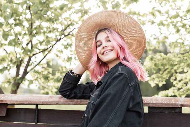Smiling young woman with pink hair in straw hat in fashionable denim jacket in beautiful summer dress is resting near wooden fence in country. Cute fashion girl with smile enjoys weekend in nature. - Photo, Image