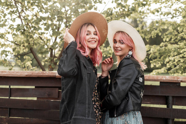 Funny smiling twin sisters with pink hair in trendy youth clothes in straw elegant hats enjoy rest outside city on sunny day. Positive girls in stylish outfit stand near wooden fence and smile happy. - Photo, Image