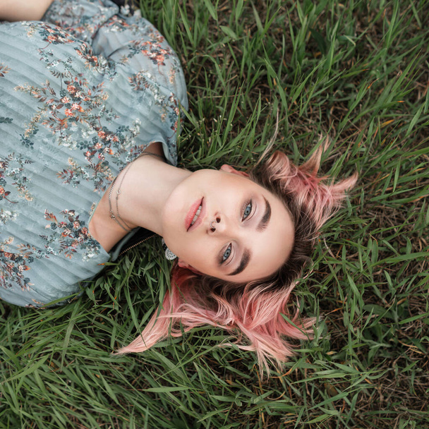 Fresh portrait charming pretty woman with clean skin with beautiful eyes with pink hair in youth elegant blue fashionable dress on green grass in field. Attractive sexy girl relaxes on lawn on nature. - Foto, afbeelding