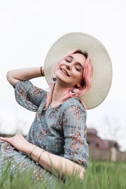 Happy fashion model mooie jonge vrouw met glimlach met gekleurd haar in elegante blauwe jurk met bloemenprint in hoed rust op groen gras in het veld buiten de stad. Glimlachend meisje in stijlvolle outfit buiten. - Foto, afbeelding