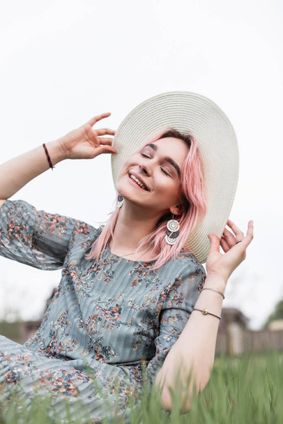 Moderne vrolijke mode jonge vrouw met glimlach met roze haar in stro witte hoed in de zomer stijlvolle jurk met bloemenprint zit op gras in het veld. Positief trendy meisje in de jeugd dragen genieten van rust op de natuur - Foto, afbeelding