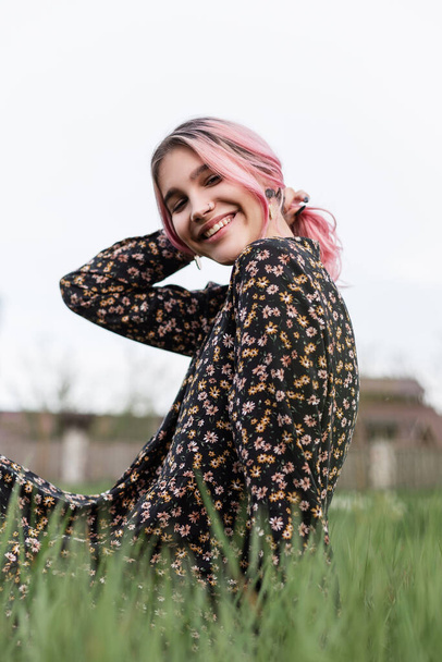 Fashion model positive beautiful woman with pink hair with lovely smile in trendy black dress with floral print resting on lawn outside city. Cute girl straightens hair and smiles. Beauty smiling lady - Photo, Image