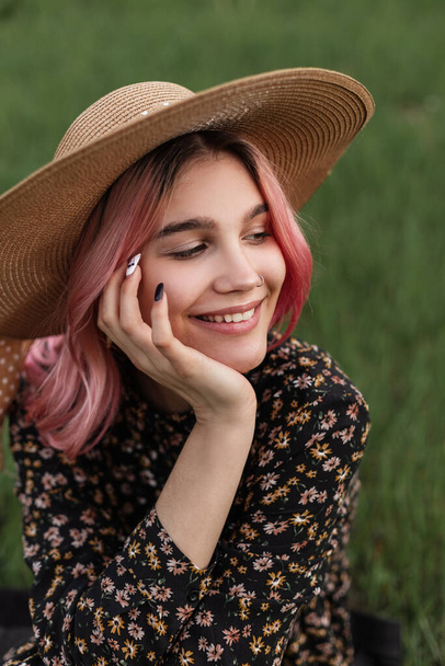 Porträt lächelnde junge feine Frau mit rosa Haaren in modischem Strohhut in stylischem schwarzen Kleid mit Blumenmuster draußen vor der Stadt. Nettes glückliches Mädchen mit schönem Lächeln ruht auf Gras auf der Natur. - Foto, Bild