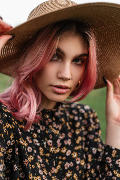 Close-up portrait attractive woman with pink hair with sexy lips with clean skin with piercing in straw fashionable hat in stylish summer black dress outdoors on nature. Beautiful elegant girl posing. - Foto, Imagen