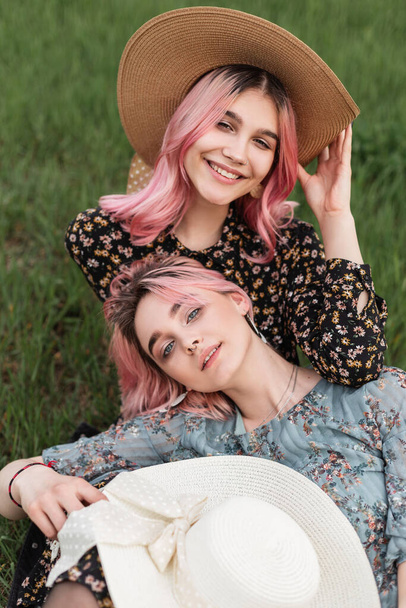Happy pretty woman with cute smile in trendy black dress in straw hat sit on grass with girlfriend in blue summer dress. Beautiful pink-haired girls in fashionable clothes rests in field outside city. - Photo, Image