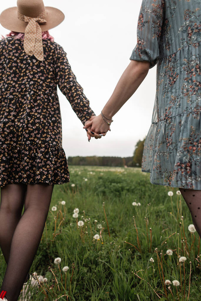 Zwei moderne Lesben in trendigen blauen und schwarzen Kleidern mit wunderschönem Blumenmuster stehen auf einem Feld zwischen grünem Gras und halten sich an den Händen. Modische Freundin Mädchen genießen Erholung im Freien. - Foto, Bild