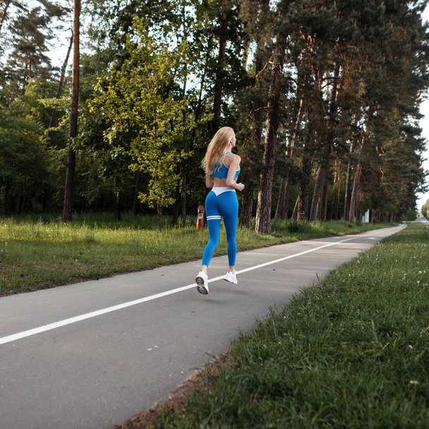 sportive beautiful young blonde girl in blue sportswear with white sneakers runs on the nature in the park - Fotografie, Obrázek