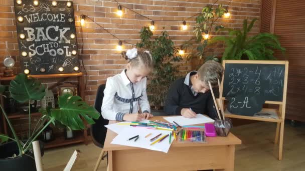 Schoolkinderen - een meisje en een jongen - zitten aan hun bureau en tekenen met gekleurde potloden tegen de achtergrond van verse bloemen en een bord met de inscriptie - Welkom! Terug naar school! - Video