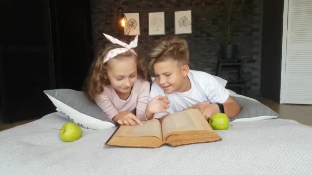Enfants - un garçon et une fille allongés sur le lit avec une grande encyclopédie et des pommes, lisent, parlent. Enseignement à domicile. - Séquence, vidéo