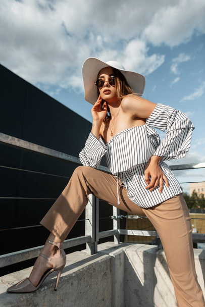 Cool stylové mladé modelky dívka s módní kulaté sluneční brýle v módní klobouk se stylovou letní top, kalhoty a boty pózovat na ulici se slunečním světlem - Fotografie, Obrázek