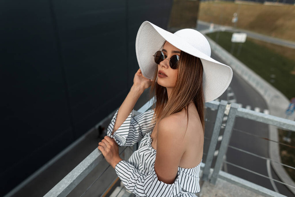 Stylish beautiful young girl in trendy summer clothes with a hat, glasses and a fashion striped top stands near a modern building in the city - Photo, Image