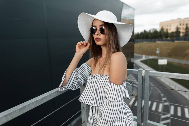 Beautiful young fashionable girl in a stylish summer hat with vintage round black sun glasses in a trendy striped top looks at the camera in the city - Фото, зображення