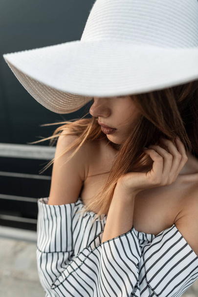 Portret van jonge mooie vrouw in modieuze witte hoed in gestreepte strandkleding nabij zwarte muur in de stad. Zomer portret van meisje een hoed covers gezicht buiten. - Foto, afbeelding