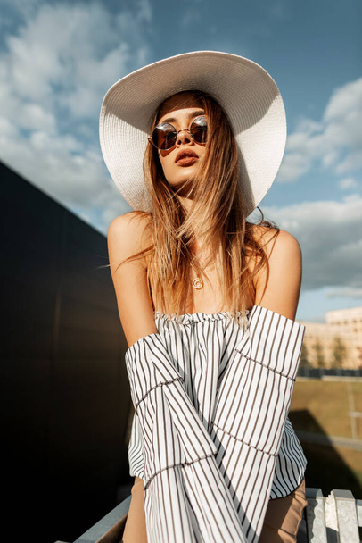 Divatos gyönyörű fiatal nő elegáns nyári ruhák szemüveggel, egy kalap és csíkos felső a városban a napfényben háttal az ég - Fotó, kép