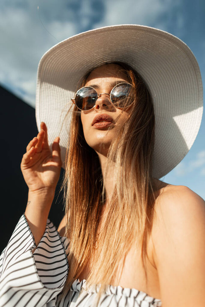 Модная красивая молодая женщина с красивыми губами в модных солнцезащитных очках с соломенной белой шляпой в полосатой блузке на пляже. Модная коллекция одежды для летнего отдыха - Фото, изображение
