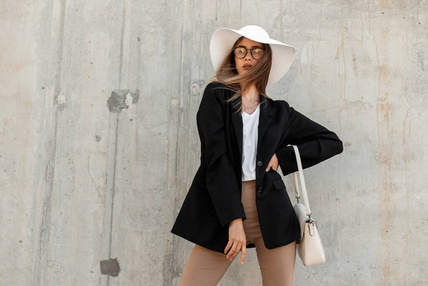 Krásná módní mladá žena s taškou v létě vintage bílý klobouk v módní černé sako v sexy top v elegantních brýlích stojí v blízkosti šedé zdi na ulici. Nenáročné pracovní oblečení. - Fotografie, Obrázek