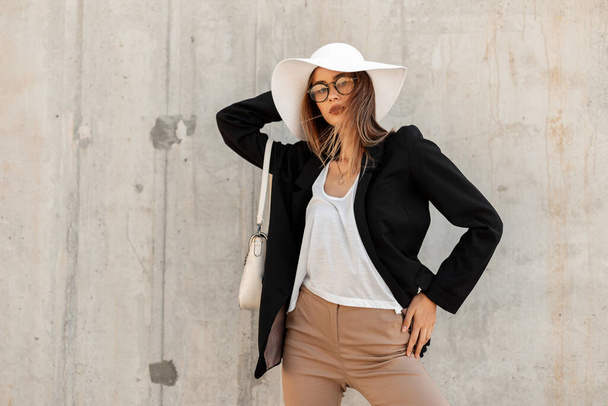 Μόδα πανέμορφη νεαρή γυναίκα το καλοκαίρι καπέλο σε κομψά γυαλιά σε μοντέρνα ασπρόμαυρα ρούχα και με δερμάτινη τσάντα θέτει σε γκρι φόντο σε εξωτερικούς χώρους. - Φωτογραφία, εικόνα
