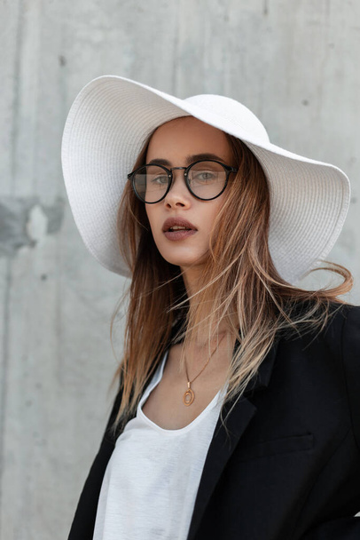 Jovem linda europeia em óculos elegantes em roupas preto-brancas na moda e chapéu de verão na praia em fundo cinza. - Foto, Imagem