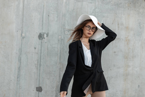 Ελκυστική σέξι νεαρή γυναίκα μοντέλο σε μοντέρνα ασπρόμαυρα ρούχα σε κομψά γυαλιά ισιώνει ψάθινο καπέλο μόδας κοντά σε γκρι κτίριο στην πόλη. Μοντέρνο επιχειρηματικό καλοκαιρινό ντύσιμο. - Φωτογραφία, εικόνα