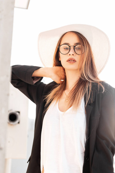 Sunny portrait pretty sexy stylish young woman in trendy glasses in fashionable black jacket in white t-shirt in straw hat outdoors. Attractive girl fashion model in youth casual wear at bright sunset - Photo, Image