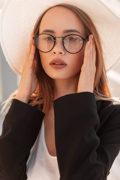 Lovely young woman in fashionable hat in stylish black blazer in white T-shirt straightens vintage glasses on bright sunny day outdoors. Gorgeous amazing girl in city. Beautiful lady and sunlight. - 写真・画像