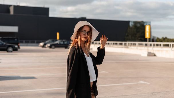 Young vogue woman in fashion black blazer and stylish top in summer beautiful hat poses outdoors in sunny day. Trendy girl model in business clothes on parking. Elegant look. - Zdjęcie, obraz