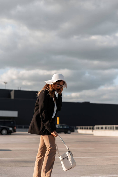 Divatos gyönyörű lány elegáns alkalmi ruhák egy zsák és egy kalap sétál végig a parkolóban a városban - Fotó, kép