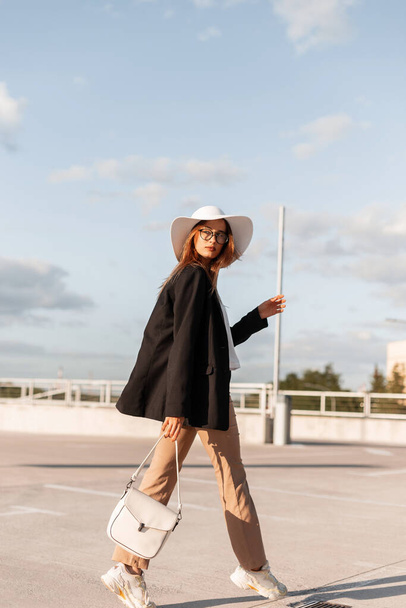 Mujer Vogue en blazer negro de moda y pantalones elegantes en sombrero elegante en zapato con bolsa de viaje al aire libre en el día soleado. Cinturón de moda en ropa de negocios en el aparcamiento. - Foto, Imagen