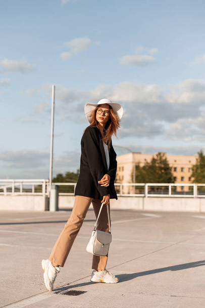 Fashionable beautiful young woman in glasses with a hat in summer stylish clothes with a fashion bag walks in the city. - Foto, Imagen