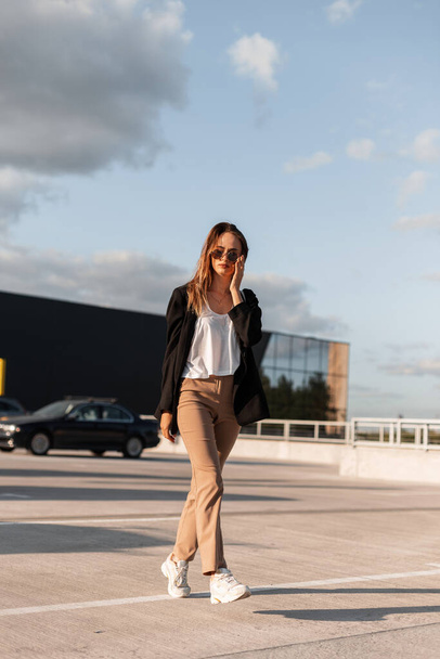 Cool young woman in stylish black jacket in vintage trousers in fashion sunglasses in top walks on asphalt on background blue sky  on sunny bright summer day. Fashionable girl travel in city car park. - Photo, image