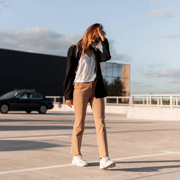 Urban young woman in black jacket in vintage trousers in fashion sunglasses in sexy top walks on asphalt and straightens hair. Beautiful girl walks in city car park on a sunny bright summer day. - Foto, Imagem