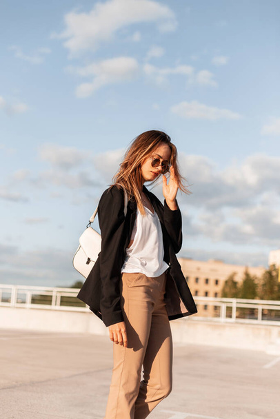 Modern beautiful woman in fashionable business clothes with bag walks in parking lot and straightens glasses. Pretty attractive girl in elegant casual dress on sunny day on background blue sky. - Zdjęcie, obraz