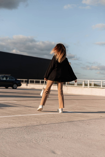 Fashionable young woman with long brown hair in stylish casual wear is dancing in an outdoor city car park on a sunny summer day. Back view. - Photo, image