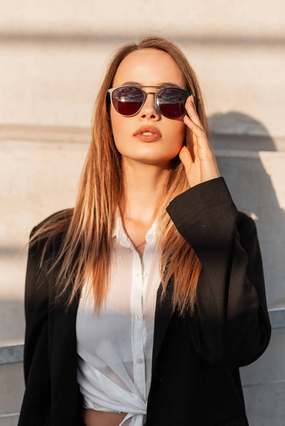 Attractive young woman in black stylish blazer straightens sunglasses and enjoys bright sunshine. Beautiful pretty urban girl in casual vintage clothes posing near building in city at sunset. - Foto, imagen
