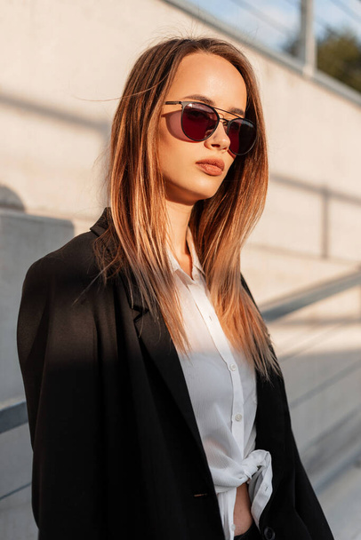 Fashionable young woman model in trendy sunglasses in fashion casual black jacket in beauty stylish white shirt near vintage wall on summer sunny day. Cool modern girl in youth clothes on sun in city. - Foto, imagen