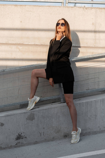 Cool mode jonge vrouw in vintage zonnebril in trendy wit shirt in zwarte modieuze jas in sneakers in de buurt van muur in de stad bij heldere zonsondergang. Stijlvol modern meisje in jeugd casual slijtage op straat. - Foto, afbeelding